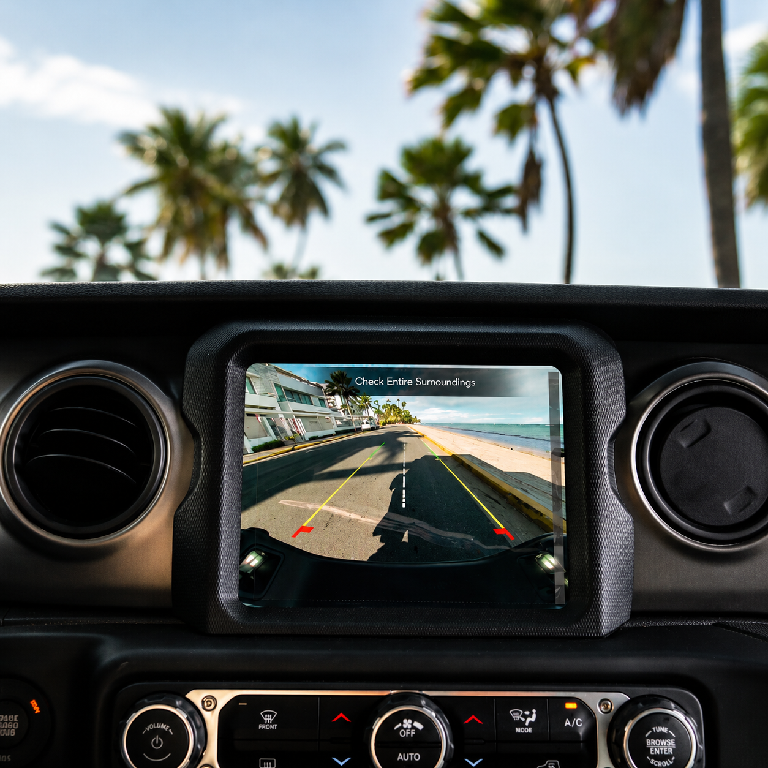 Jeep dashboard showing the backup camera view of a coastal road with palm trees
